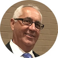 Smiling man wearing glasses and a suit, standing against a neutral-colored background. Represents a professional context.